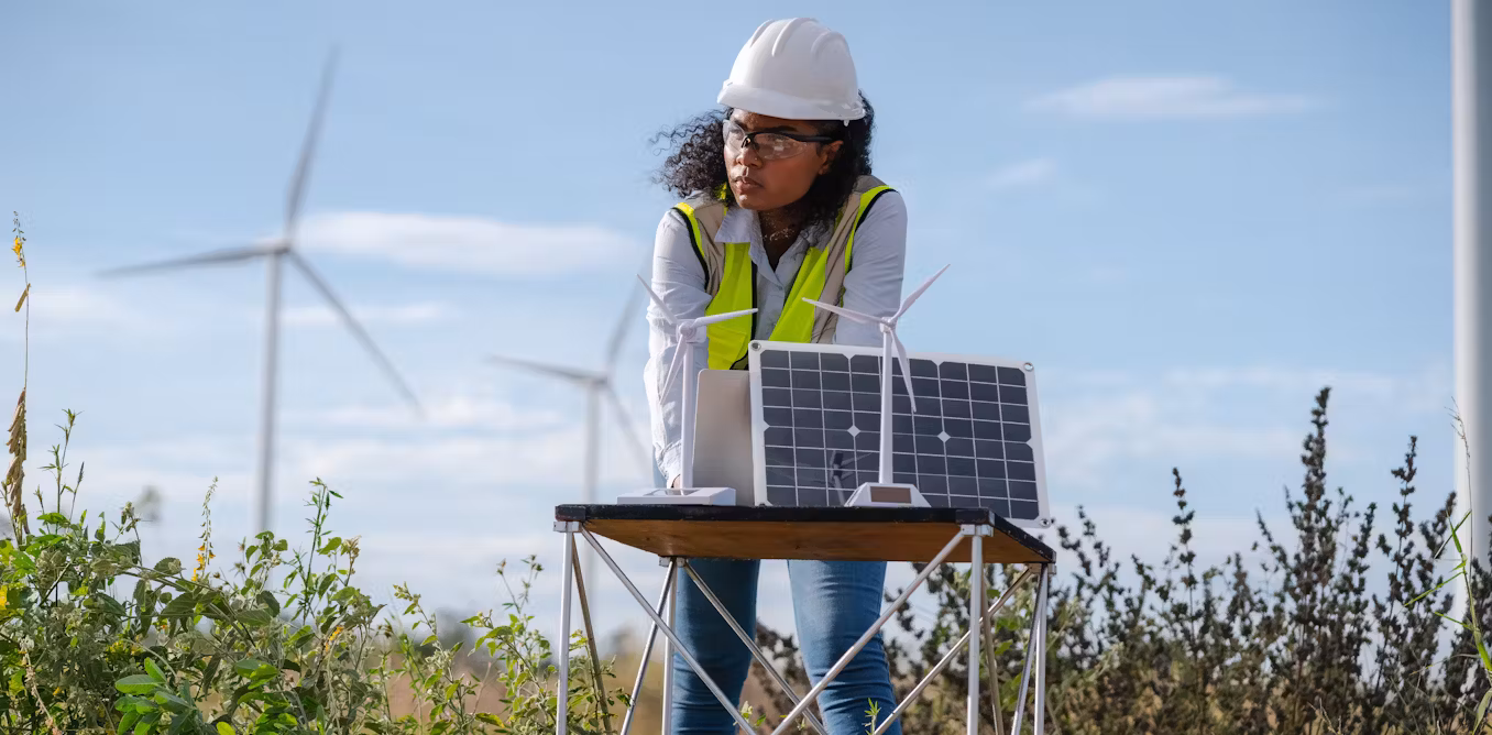 Woman in safety apparel standing behind a solar panel and model wind mills.